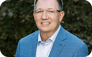 Man in blue plaid blazer and glasses smiling outdoors against green foliage