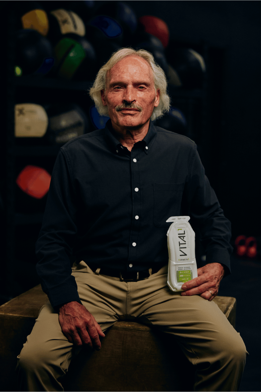Elderly man with white hair holding a "VITAL1" drink bottle, seated in a dark gym with colorful boxing gloves behind him