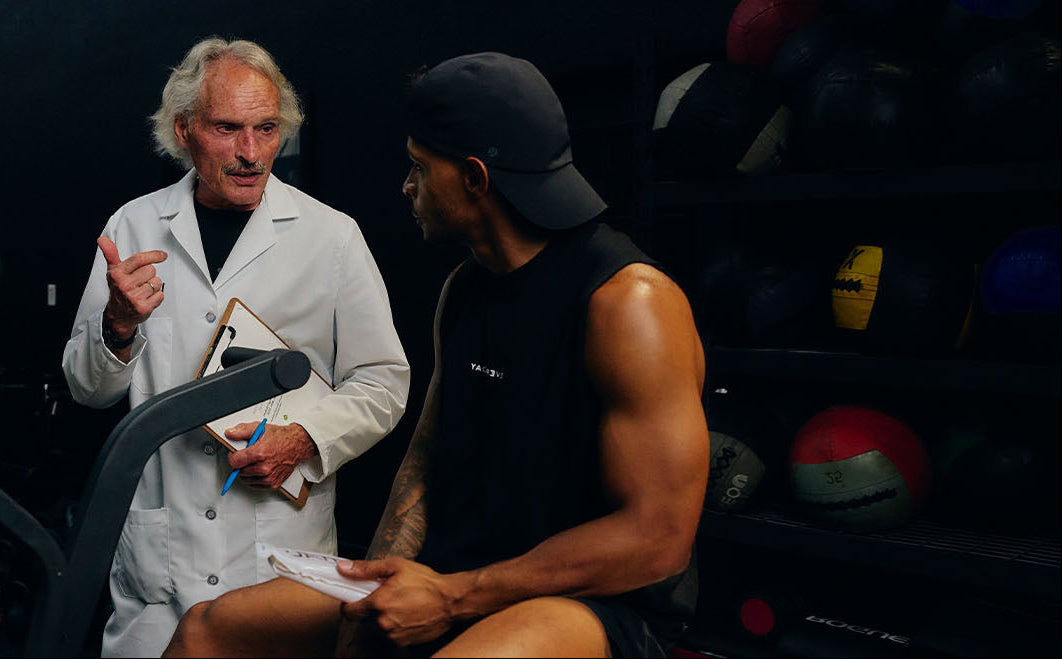 A white-coated doctor with clipboard consults a muscular athlete in a dark gym with medicine balls