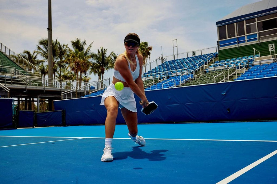 Woman in white athletic outfit playing pickleball on blue outdoor court with palm trees and stadium seating behind her