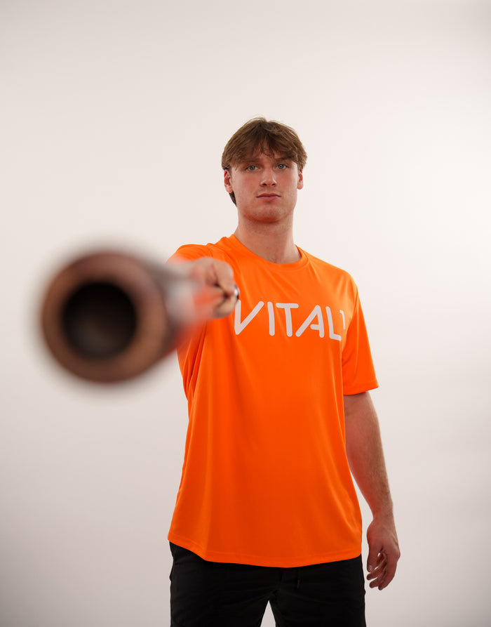 Young man in orange "VITALI" shirt pointing dumbbell toward camera against white background