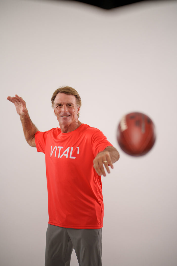 Man in red "VITAL1" shirt throwing a football in a white studio setting