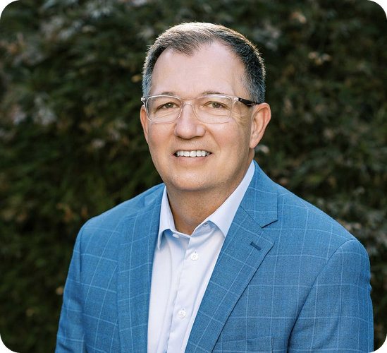 Man in blue plaid blazer and glasses smiling outdoors with green foliage background