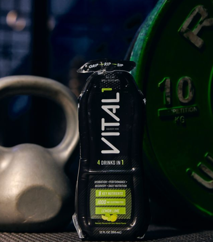 VITAL1 black lemon lime sports drink bottle, "4 Drinks In 1," beside kettlebell and 10kg weight plate
