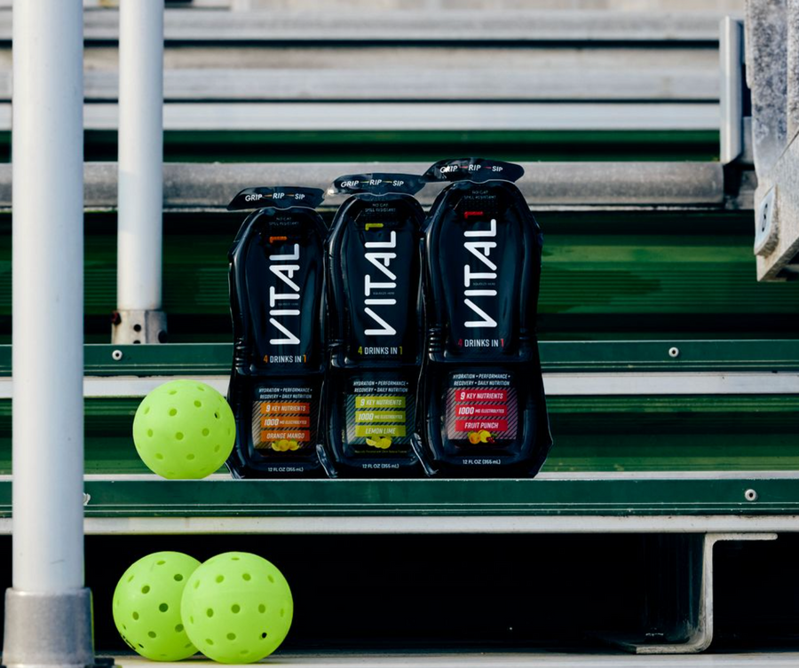 Three black VITAL "4 Drinks in 1" sport drink pouches in Orange Mango, Lemon Lime, and Fruit Punch flavors on green bleachers with yellow pickleballs nearby.