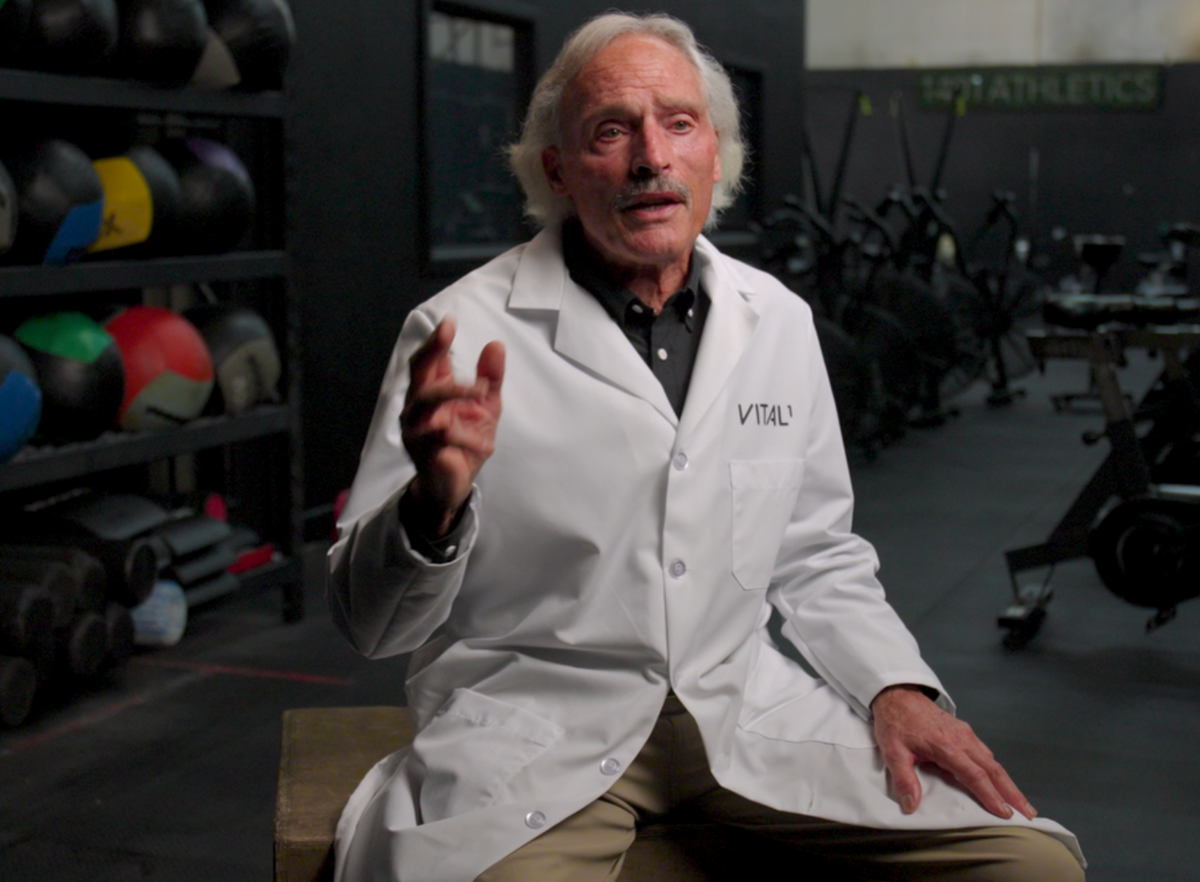 Elderly man in white "VITAL" lab coat seated in gym, gesturing while speaking, medicine balls visible behind him