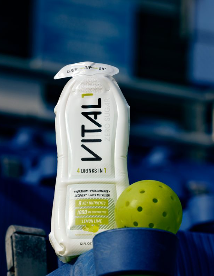 White VITAL1 lemon lime sports drink pouch beside a yellow pickleball, blue court background