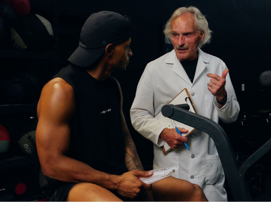 Muscular athlete in black tank consults white-coated doctor holding clipboard in dark gym