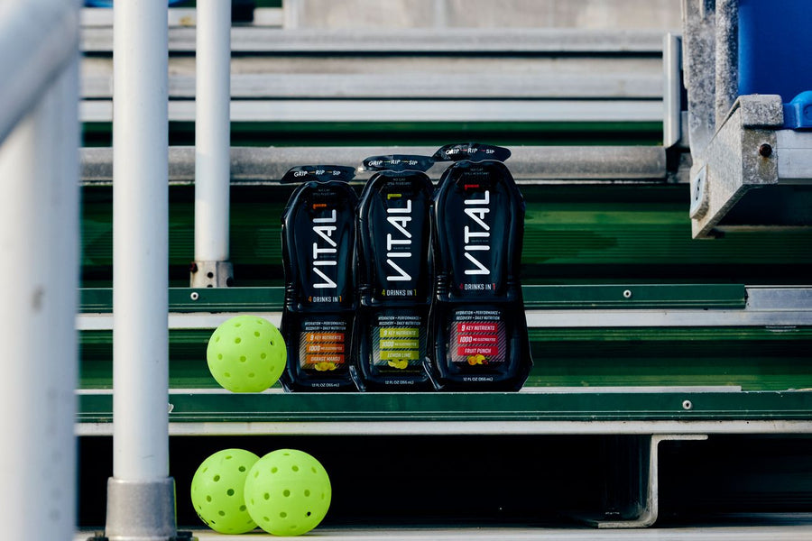 Three black VITAL "4 Drinks In 1" sports drink pouches in Orange Mango, Lemon Lime, and Fruit Punch flavors on bleacher steps beside neon yellow pickleballs